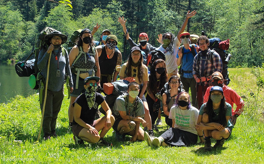 A group of people stand, squat, and sit on a patch of grass in front of trees.